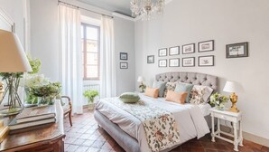 Une chambre à coucher avec un grand lit, un lustre, une fenêtre avec des rideaux et des tableaux encadrés accrochés au mur.