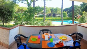 Une table ronde recouverte d’une nappe noire, entourée de quatre chaises noires dotées de coussins bleus, est placée sur un balcon donnant sur une piscine et une végétation luxuriante.