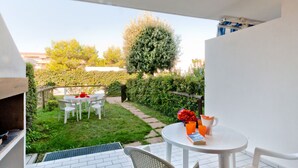 A patio with a grill, a round table with chairs, and a view of a garden with a tree and a hedge. - Villaggio Maja by Interhome (Bibione)