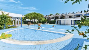 A swimming pool area with a tiled deck, a covered seating area, and a building with multiple windows in the background. - Villaggio Maja by Interhome (Bibione)