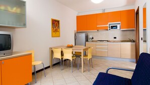 A compact kitchen with orange cabinets, a microwave, and a small refrigerator. There is a dining table with chairs and a television on a stand. - Villa Angela by Interhome (Bibione)
