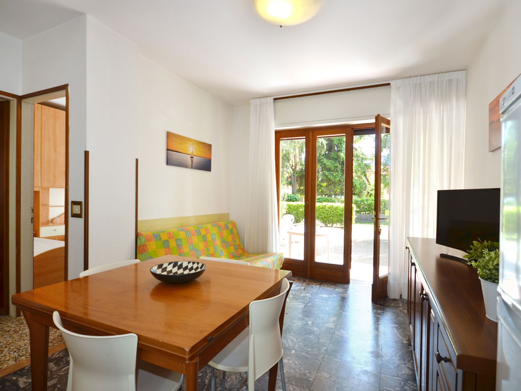 Una habitación con una mesa de madera, sillas blancas, un sofá colorido, un tazón sobre la mesa y un televisor en un mueble.