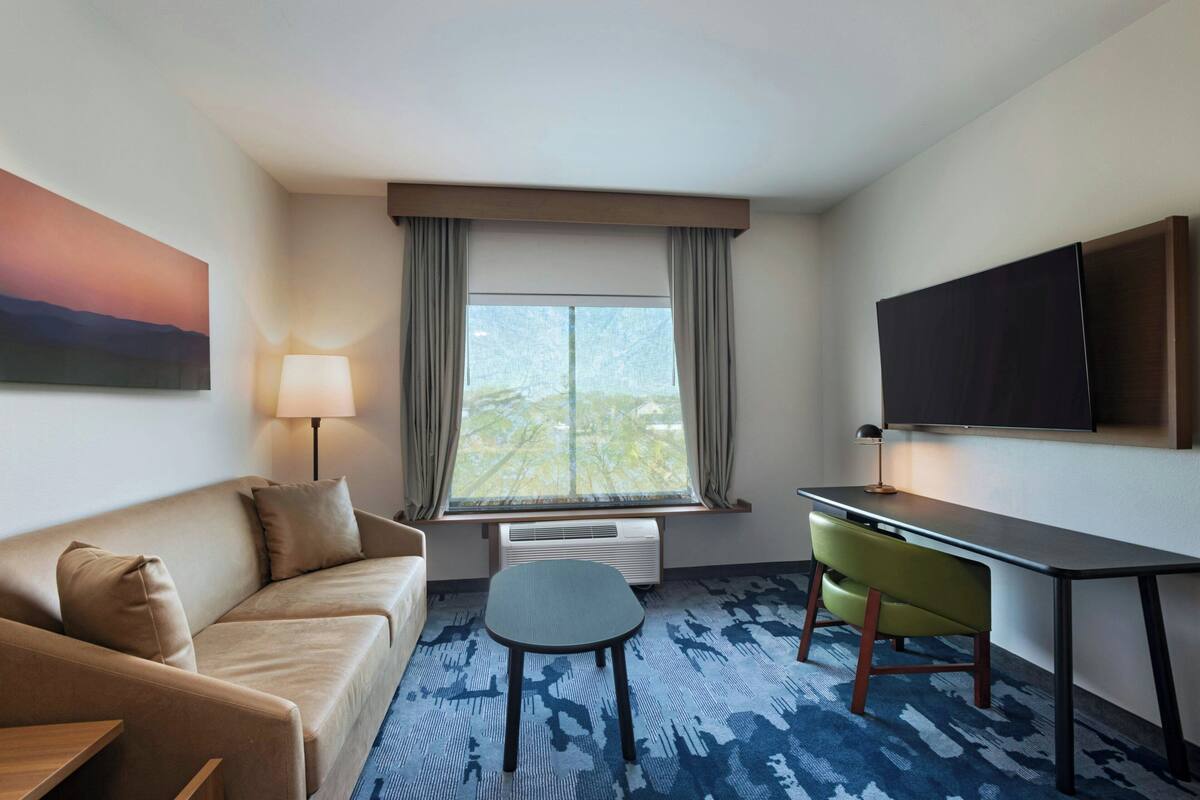 A hotel room with a sofa, a small table, a chair, a TV, and a window with curtains.