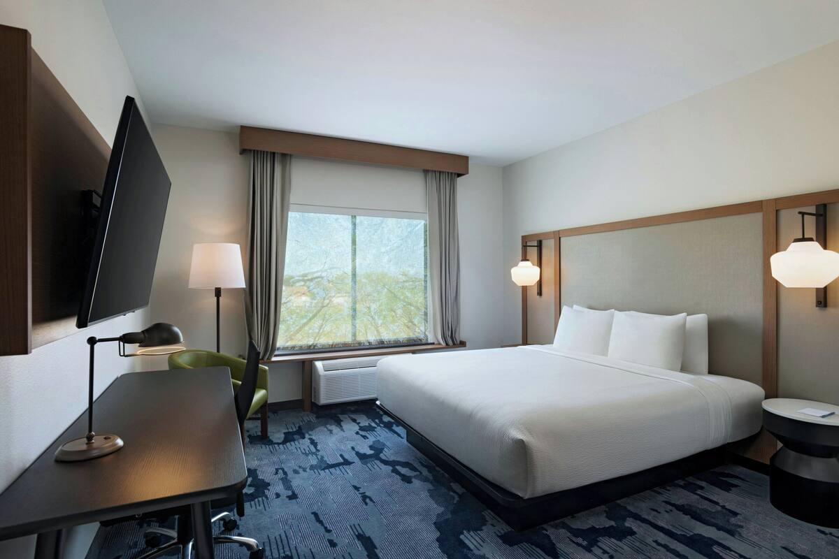 A hotel room with a large bed, a desk with a chair, a flat-screen TV, a window with curtains, and a round table with a lamp.