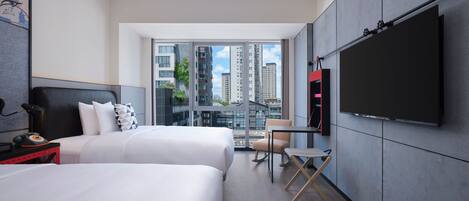 A modern hotel room with two beds, a TV, a desk, and a view of the cityscape.