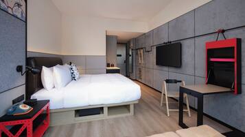 A modern hotel room with a bed, bedside table, chair, TV, and a small desk.