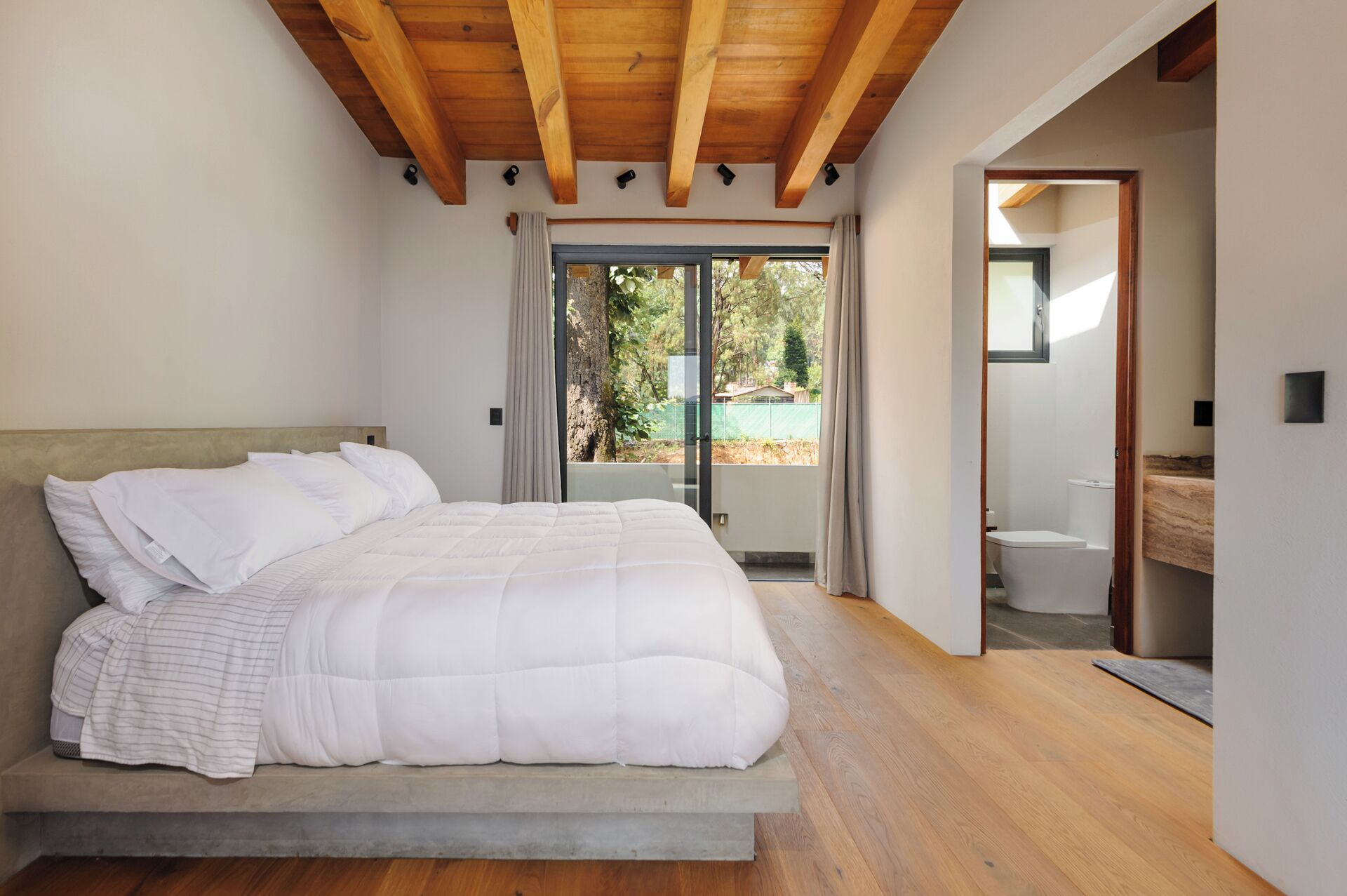 A bedroom with a large bed, wooden ceiling beams, a sliding glass door to a balcony, and a visible bathroom.