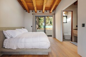 Un dormitorio con una cama grande, vigas de techo de madera, un ventanal corredero a un balcón y un baño a la vista.