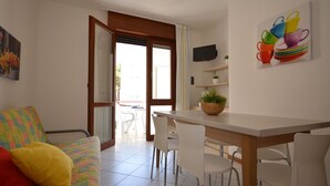 A modern living room with a colorful sofa, a dining table with chairs, a TV mounted on the wall, and a balcony with a view. - Torcello by Interhome (Bibione)