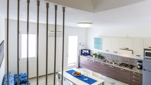 Une cuisine moderne avec des armoires blanches, un comptoir en granit et des appareils en acier inoxydable. On y trouve un coin repas avec une table blanche et un set de table bleu, ainsi qu’un canapé bleu à motifs.