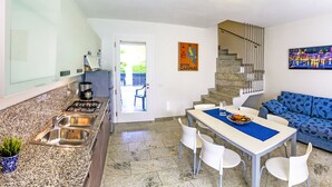 Une cuisine moderne avec des comptoirs en granit, un évier et une cuisinière. On y trouve un coin repas avec une table et des chaises, ainsi qu’un canapé bleu à motifs. Un escalier mène à l’étage supérieur.