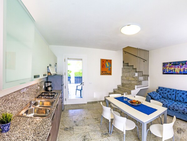 A modern kitchen with granite countertops, a sink, and a stove. There is a dining area with a table and chairs, and a blue patterned sofa. A staircase leads to an upper level.