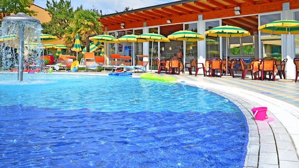 Zona de piscina con sillones de descanso y sombrillas, fuente de agua y área de estar cubierta.