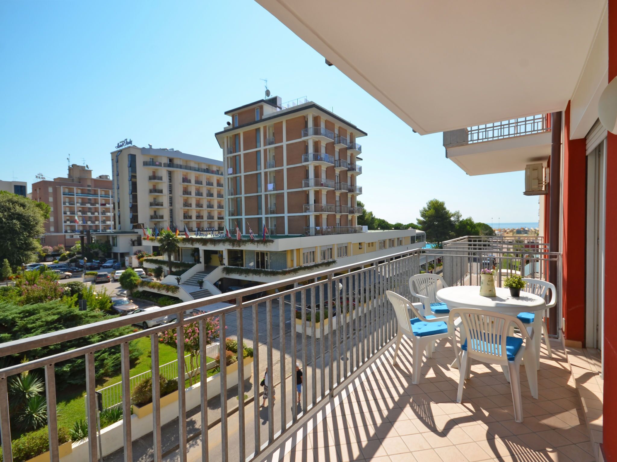A balcony with a table and chairs, overlooking a courtyard with greenery and a building with multiple balconies.