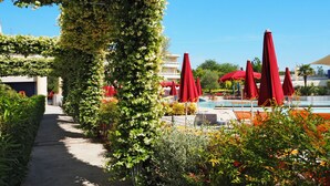 Ein mit roten Sonnenschirmen gesäumter Weg führt zu einem Poolbereich, umgeben von üppiger Vegetation und blauem Himmel.