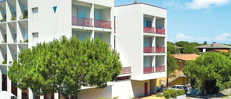 A modern multi-story building with balconies, a parking area, and surrounding greenery.
