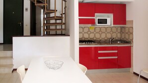 Une cuisine avec des meubles de couleur rouge, un plan de travail blanc et un four à micro-ondes. On y trouve une table à manger recouverte d’une nappe blanche et un saladier en métal. Un escalier avec des rampes en bois est visible à l’arrière-plan.