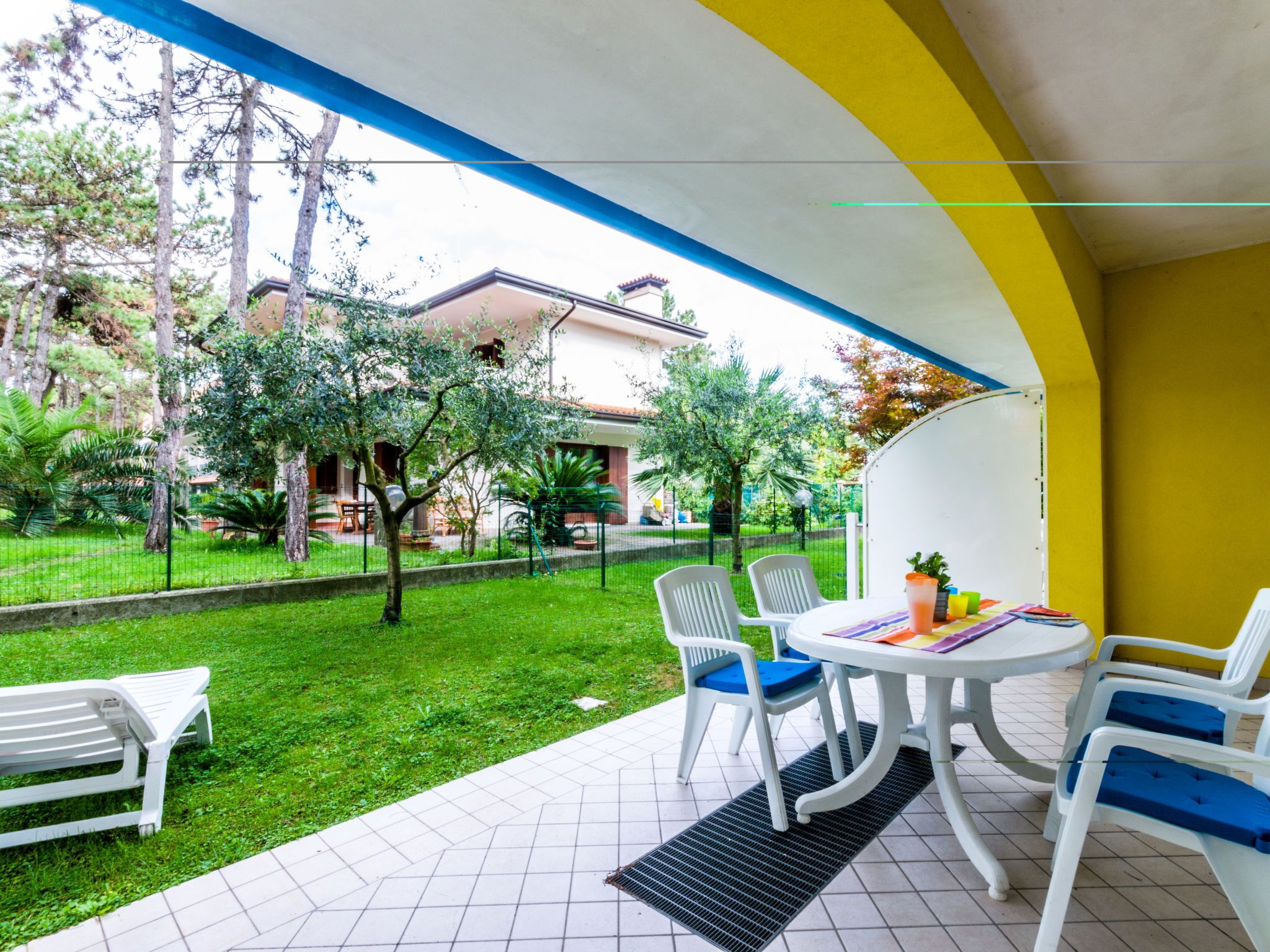 Un espace patio avec une table et des chaises blanches, un banc, et une vue sur un jardin avec des arbres et un bâtiment en arrière-plan.