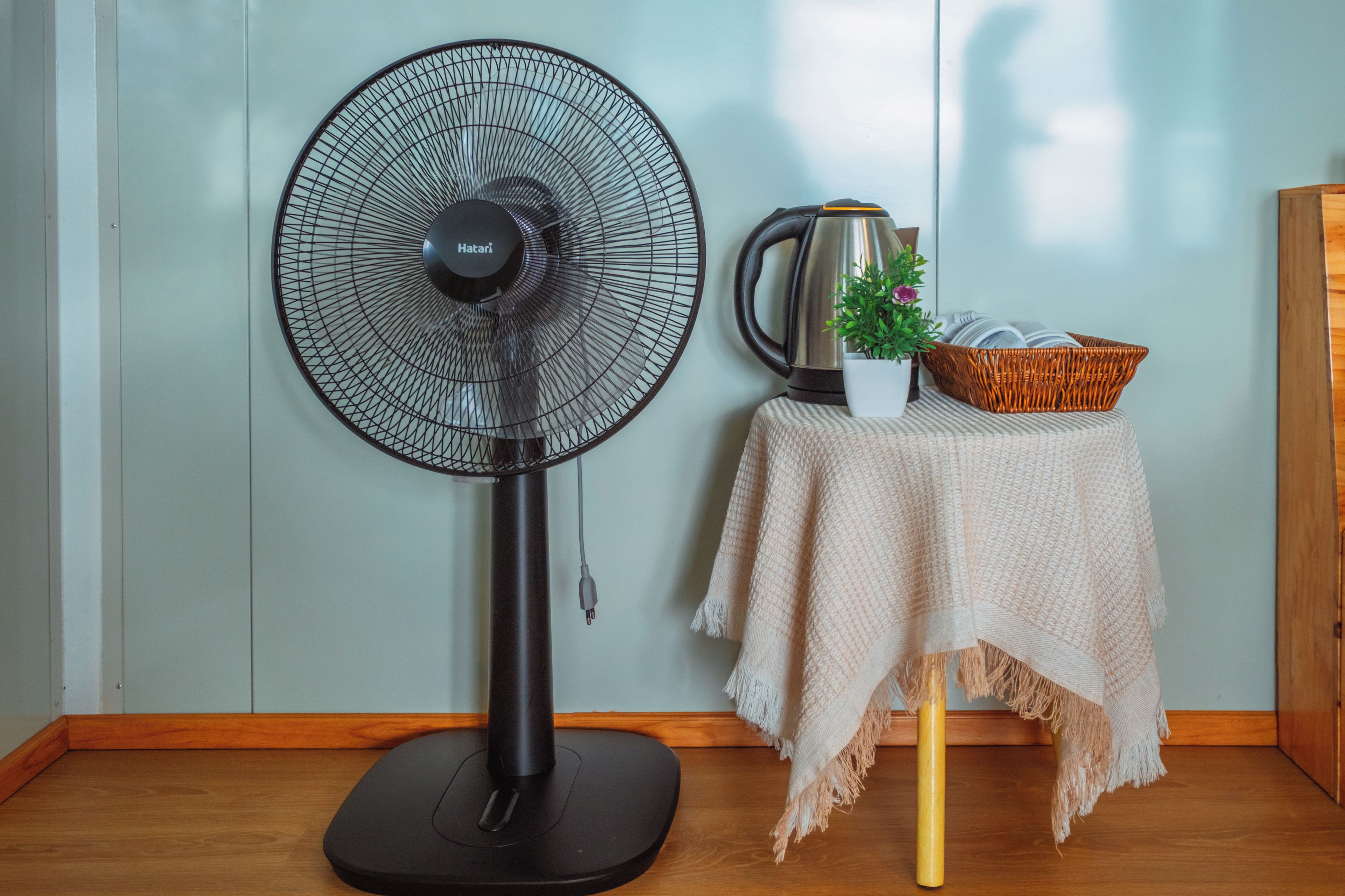 Una habitación con un ventilador de pie, un hervidor, una cesta con libros y una planta pequeña sobre una mesa.