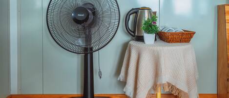 Una habitación con un ventilador de pie, un hervidor, una cesta con libros y una planta pequeña sobre una mesa.