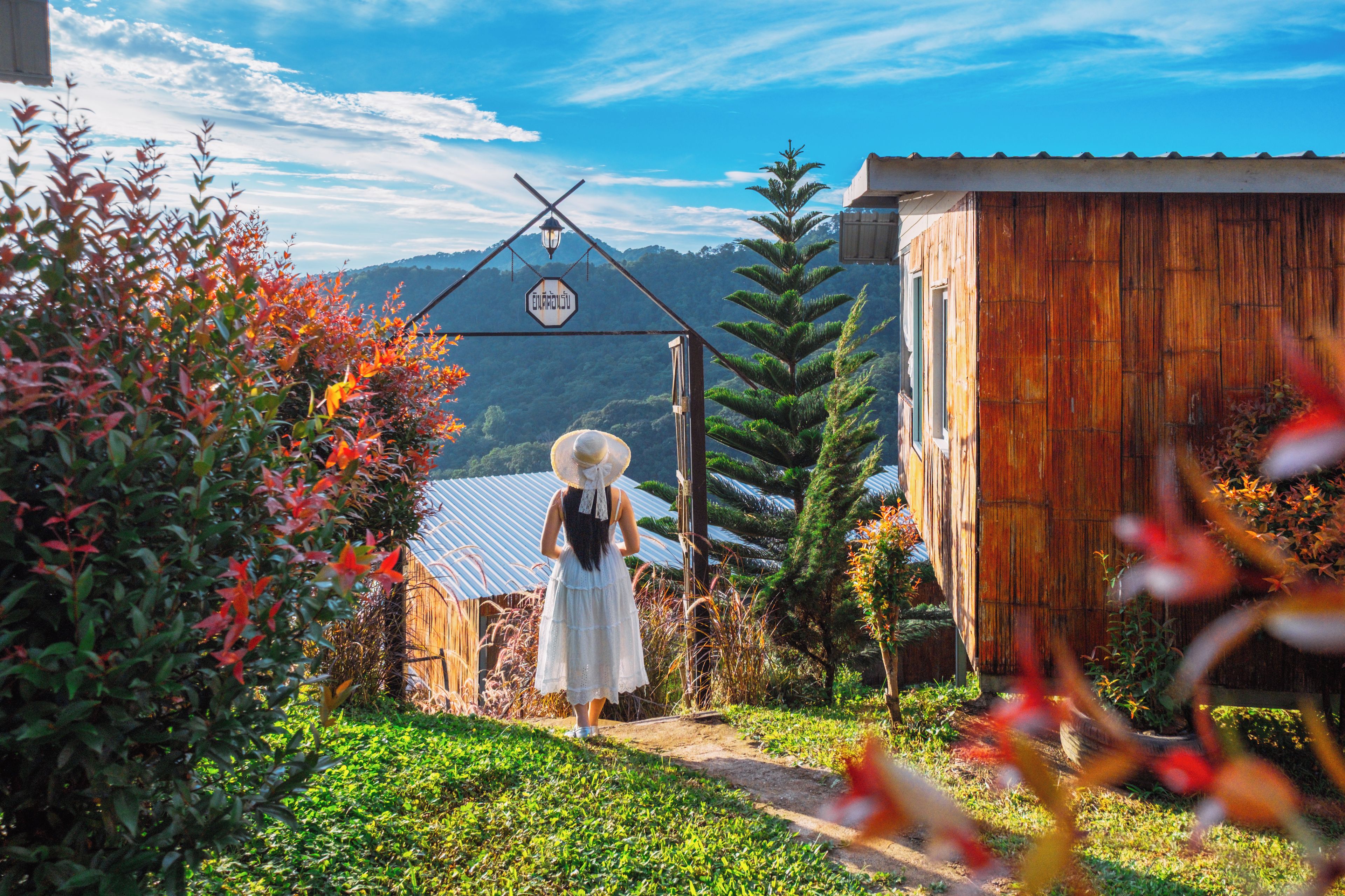 A woman in a white dress and straw hat walking towards a wooden structure with a sign above it.
