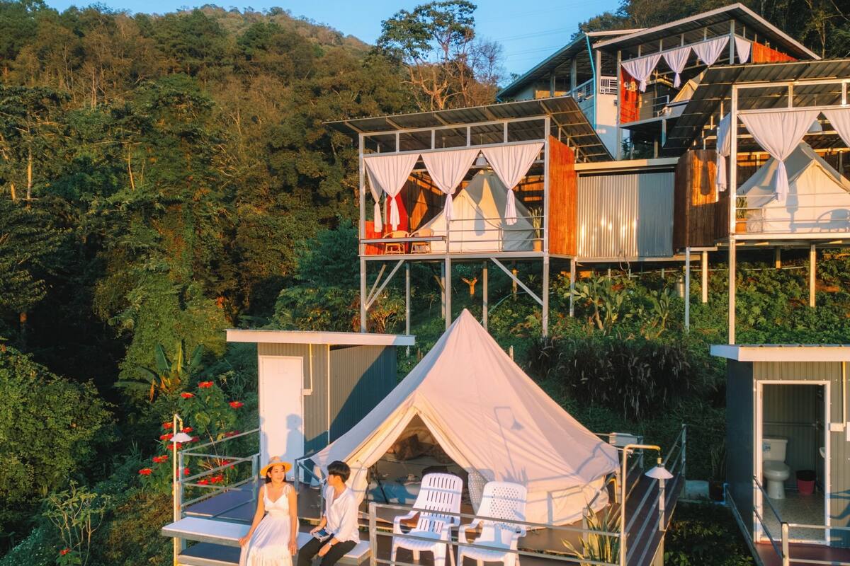 A multi-level outdoor accommodation with separate tents and cabins, surrounded by lush greenery.