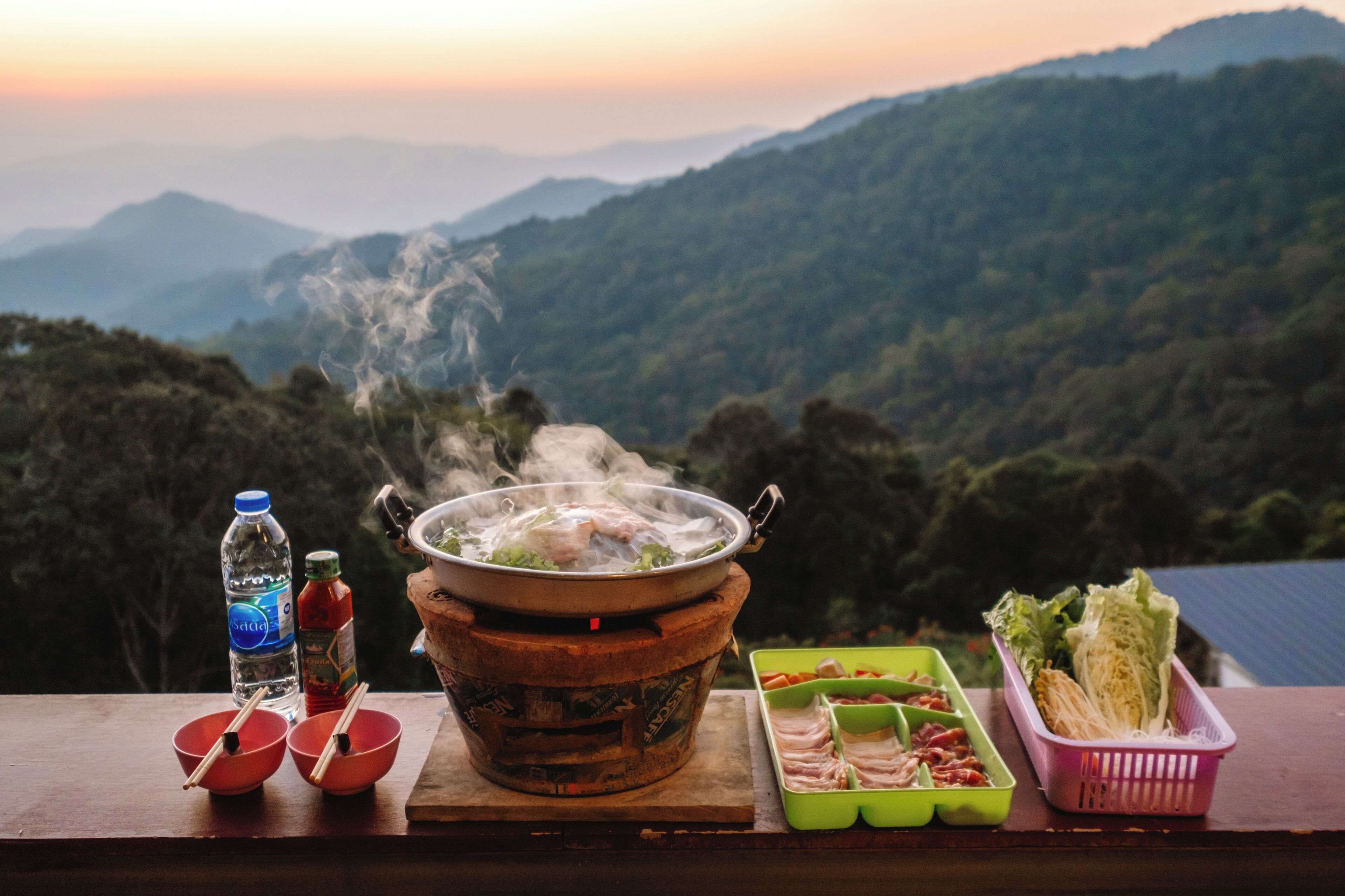 Una mesa con un recipiente de comida, palillos, una botella de agua y una bandeja verde con alimentos, con montañas al fondo.
