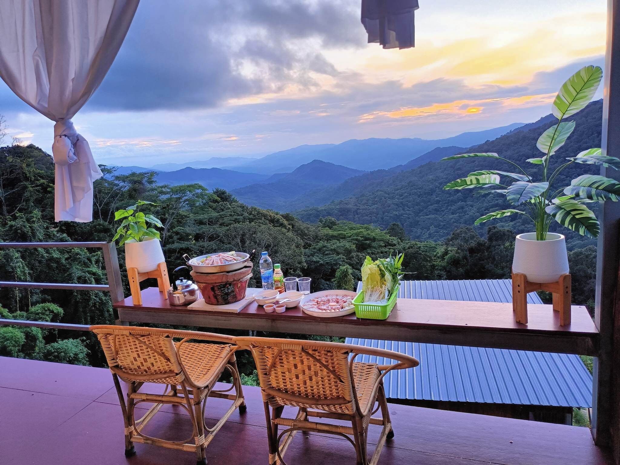Una mesa de comedor con comida y bebidas, dos sillas de mimbre y vistas a montañas.