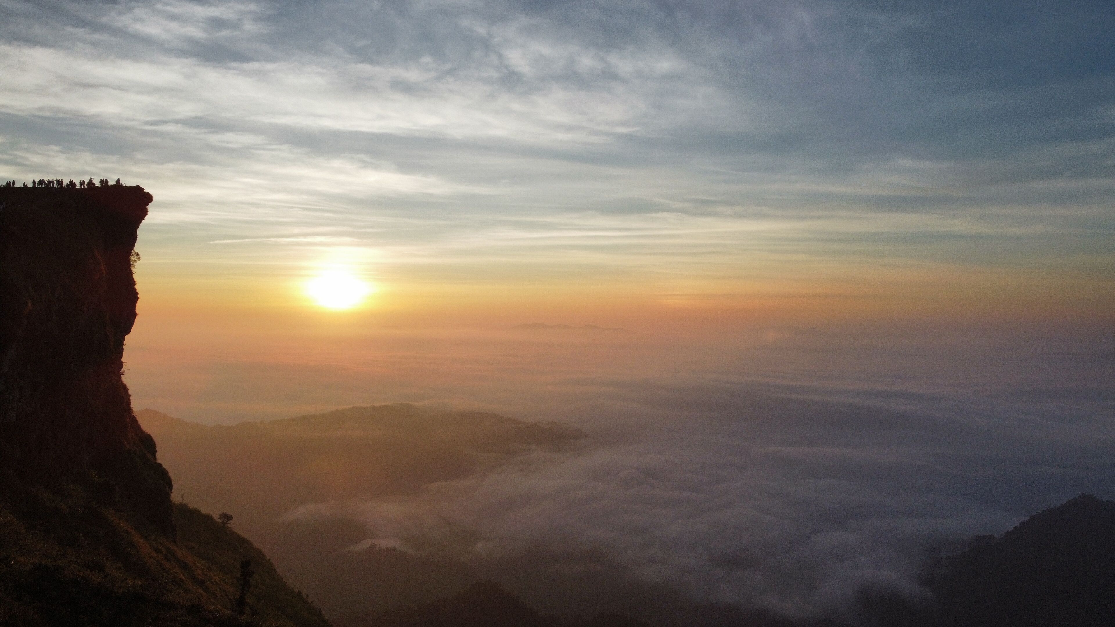A sunrise over a mountainous landscape with a cliff on the left and clouds below.
