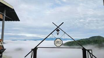 A rustic wooden structure with a metal roof, a sign with Thai script, and a view of a forested mountain range above a cloud layer.