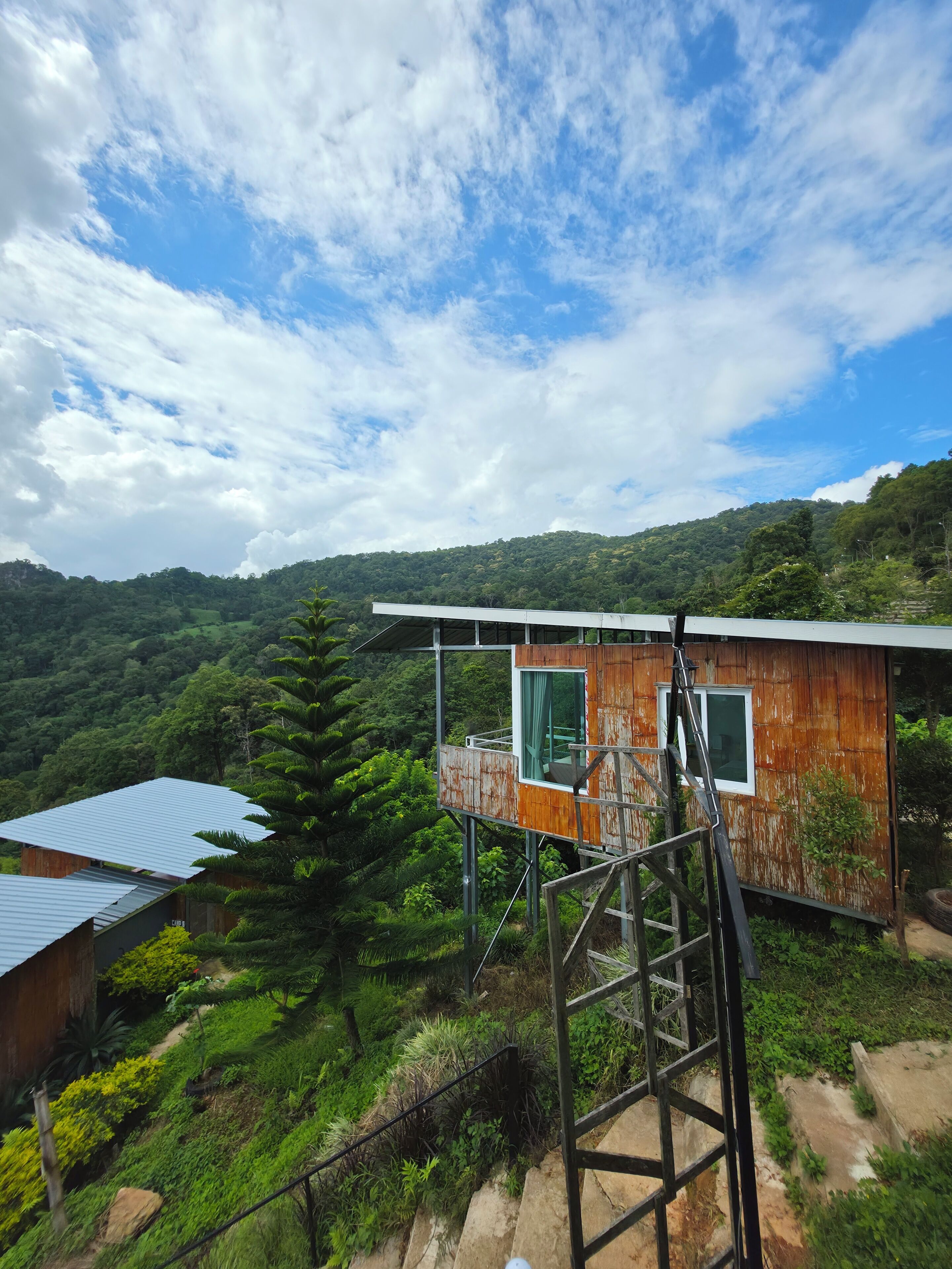 Ein Baumhaus mit einer Metalltreppe, umgeben von üppiger Vegetation und einer Berglandschaft im Hintergrund.