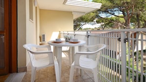 Un balcon avec une table ronde et deux chaises, une balustrade blanche, et une vue sur les arbres et les bâtiments en arrière-plan.