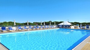 Een zwembad met blauw water, omringd door ligbedden en parasols, met een heldere lucht en groen in de achtergrond.