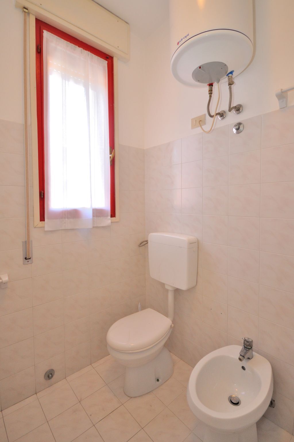 Bagno con WC, bidet e finestra con cornice rossa e tende bianche.