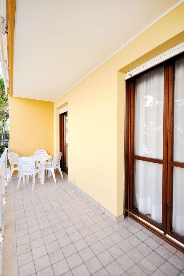A balcony with a tiled floor, a white table and chairs, and a large window with curtains. - Pleione by Interhome (Bibione)