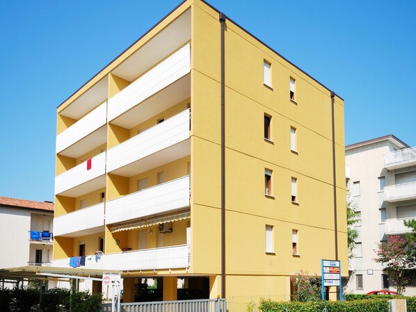 A yellow multi-story building with balconies, a visible ground floor entrance, and a signboard with various advertisements. - Pleione by Interhome (Bibione)