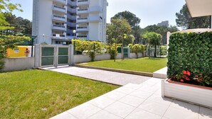 Un edificio residencial con entrada porticada, césped bien cuidado y un seto con flores rojas.
