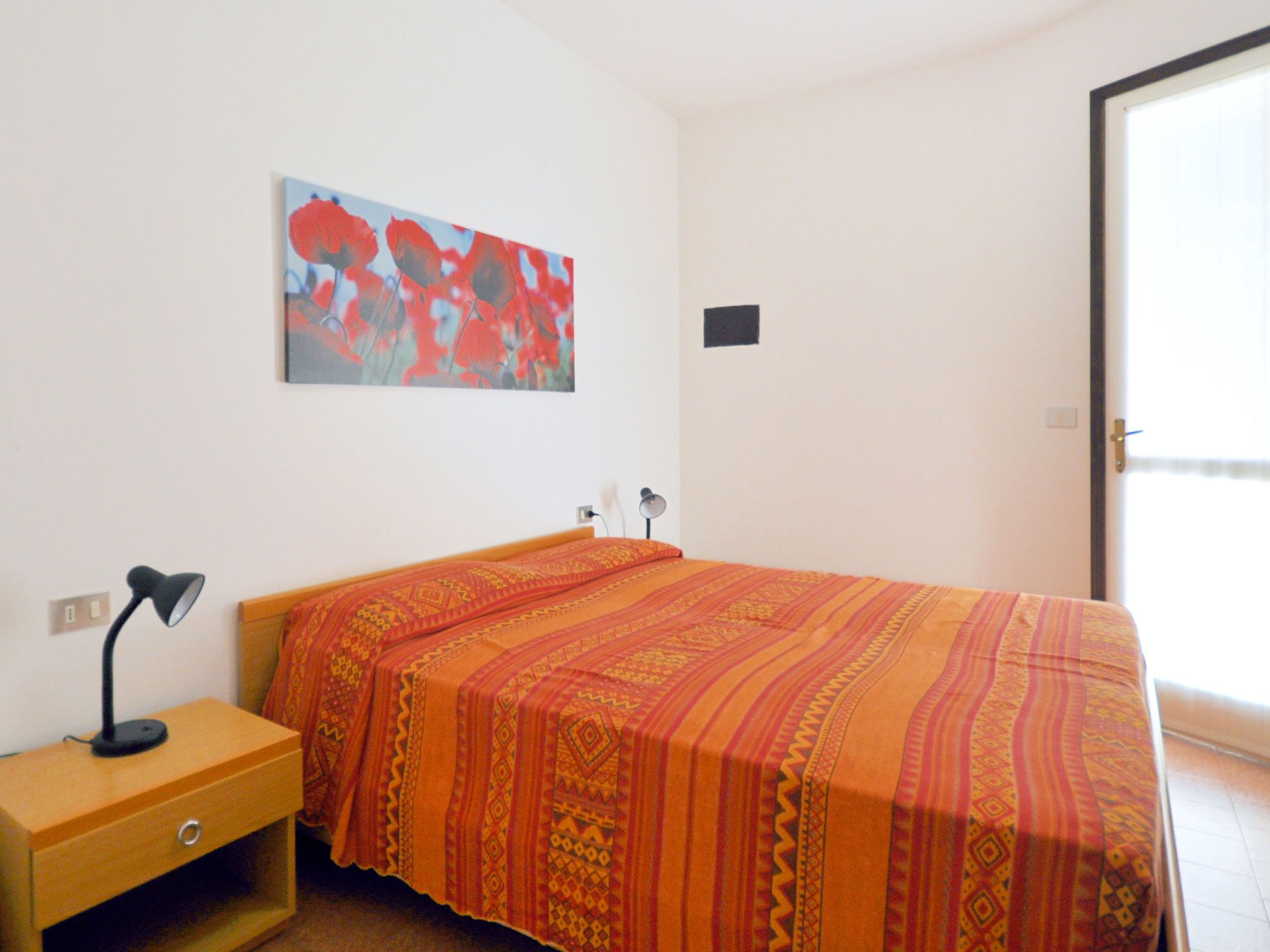 Quarto com cama, mesa de cabeceira, abajur, tomada na parede e porta.