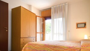 A bedroom with a wooden wardrobe, a bed with a colorful quilt, a window with curtains, and a wall-mounted picture frame. - Boschetto by Interhome (Bibione)