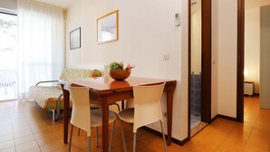 A dining area with a wooden table, four chairs, a colorful sofa, and a window with curtains. - Apartment Erica by Interhome (Bibione)