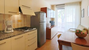 A compact kitchen with a gas stove, refrigerator, and a coffee maker. There is a dining area with a table and chairs, and a balcony with a view. - Apartment Erica by Interhome (Bibione)