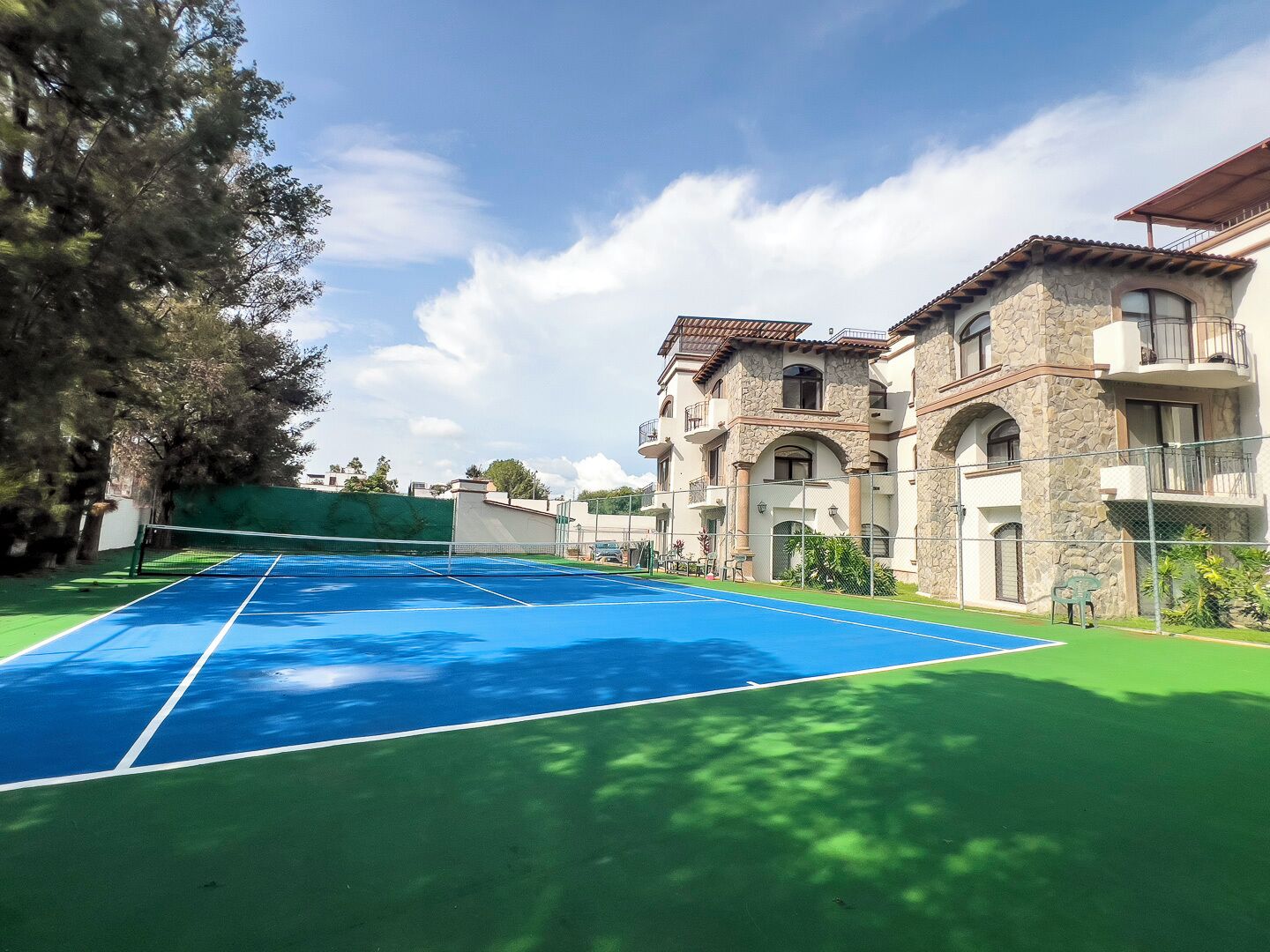 Un court de tennis avec un filet, entouré d’une zone verdoyante et d’un bâtiment dont la façade est en pierre et en stuc.