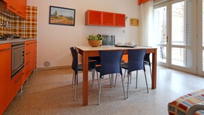 A compact kitchen with orange cabinets, a dining table with four chairs, and a window with white shutters. - Villa Verde by Interhome (Bibione)