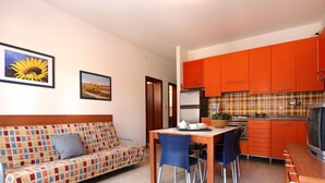 A compact living space with a sofa, a dining table with chairs, a kitchenette with orange cabinets, and two framed pictures on the wall. - Villa Verde by Interhome (Bibione)