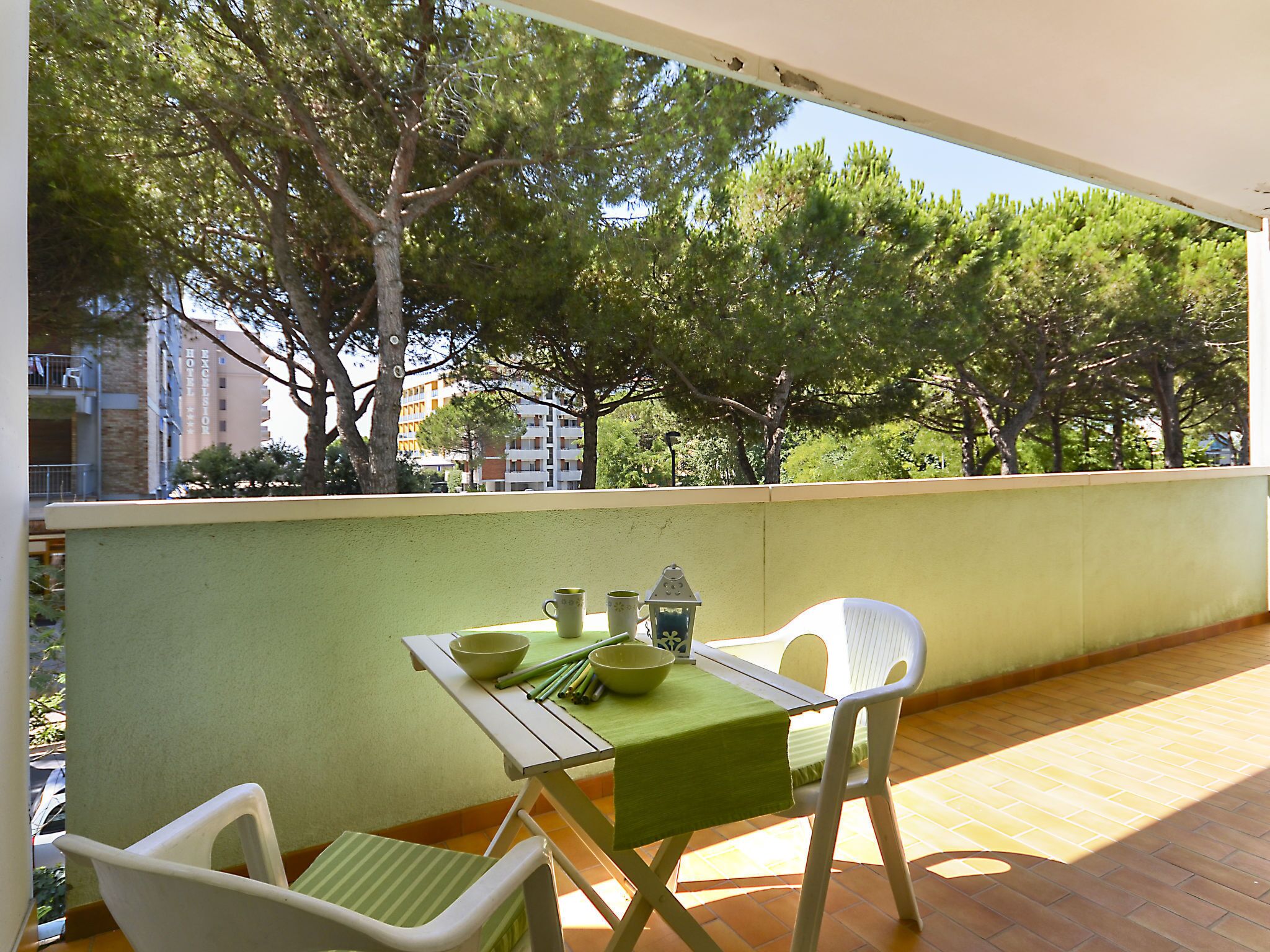A balcony with a table set for two, overlooking a tree-lined area with buildings in the background.