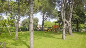 A playground with a slide and climbing structures in a grassy park area surrounded by trees. - Villaggio Splendido by Interhome (Bibione)
