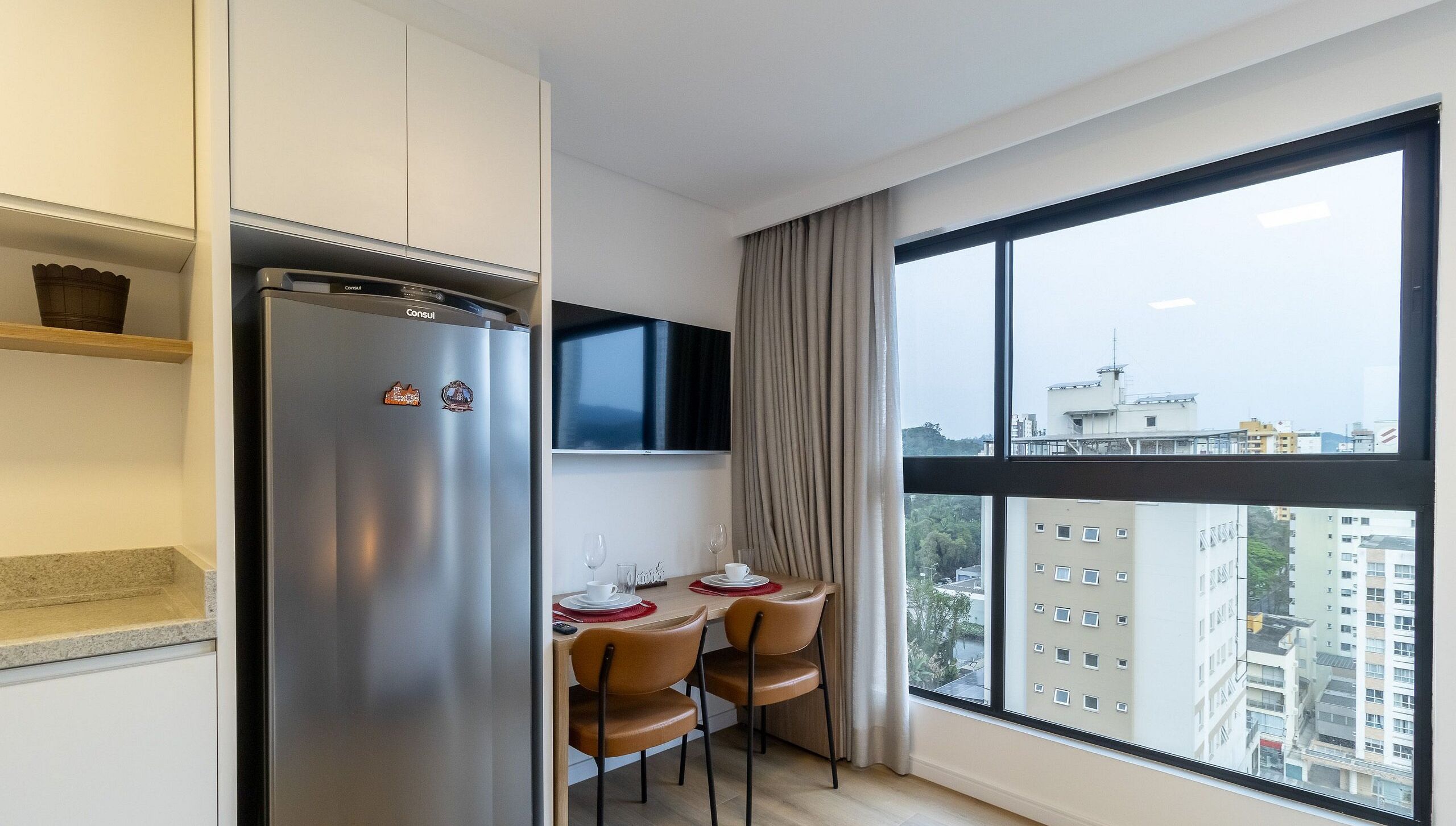A modern kitchen with a stainless steel refrigerator, a flat-screen TV mounted above the counter, and a dining area with two chairs and a table set for two.