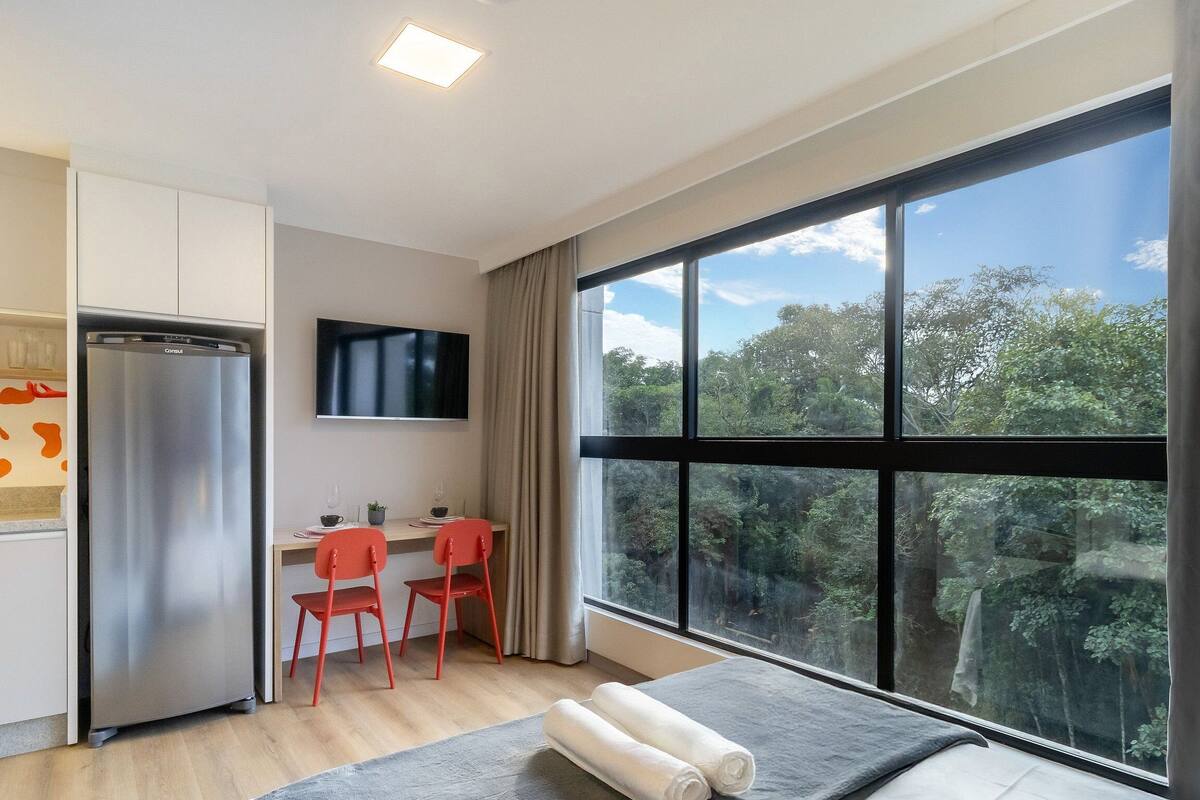 A modern kitchen with a stainless steel refrigerator, a dining area with two red chairs, and a large window offering a view of a forest.