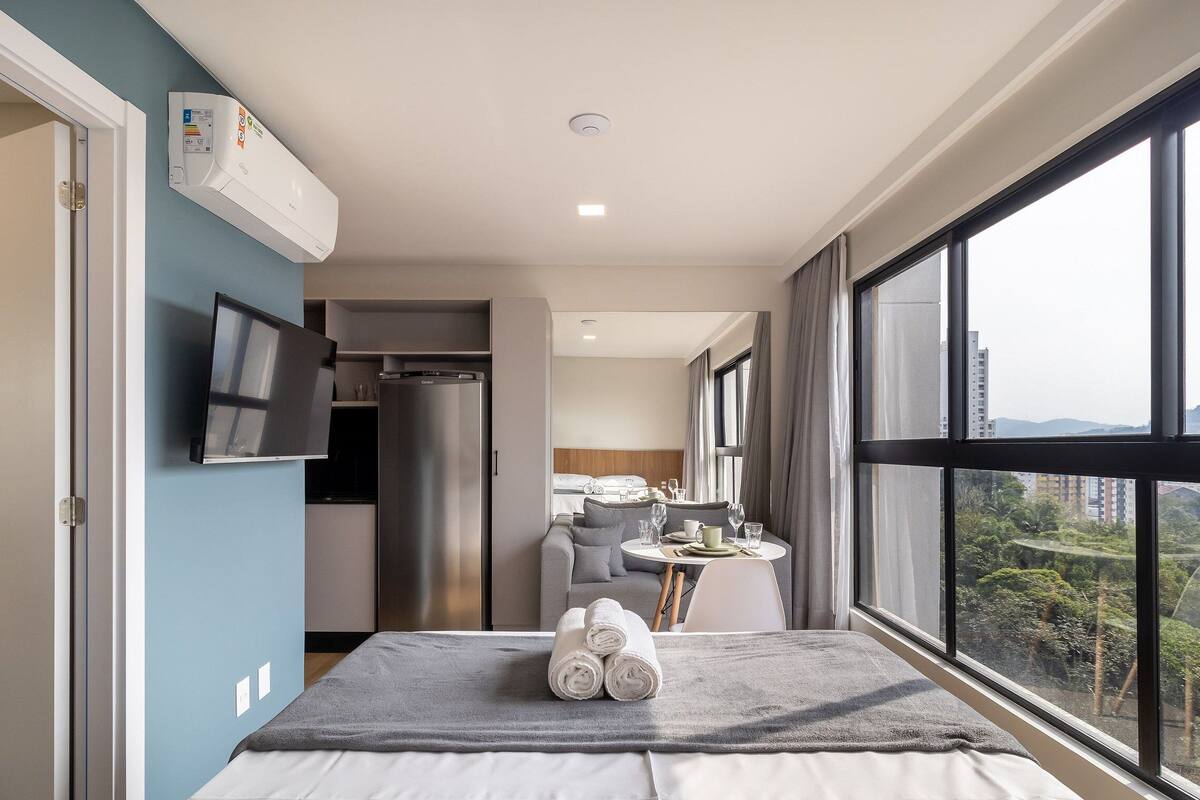 A modern hotel room with a bed, a TV, a kitchenette, and a large window with a city view.