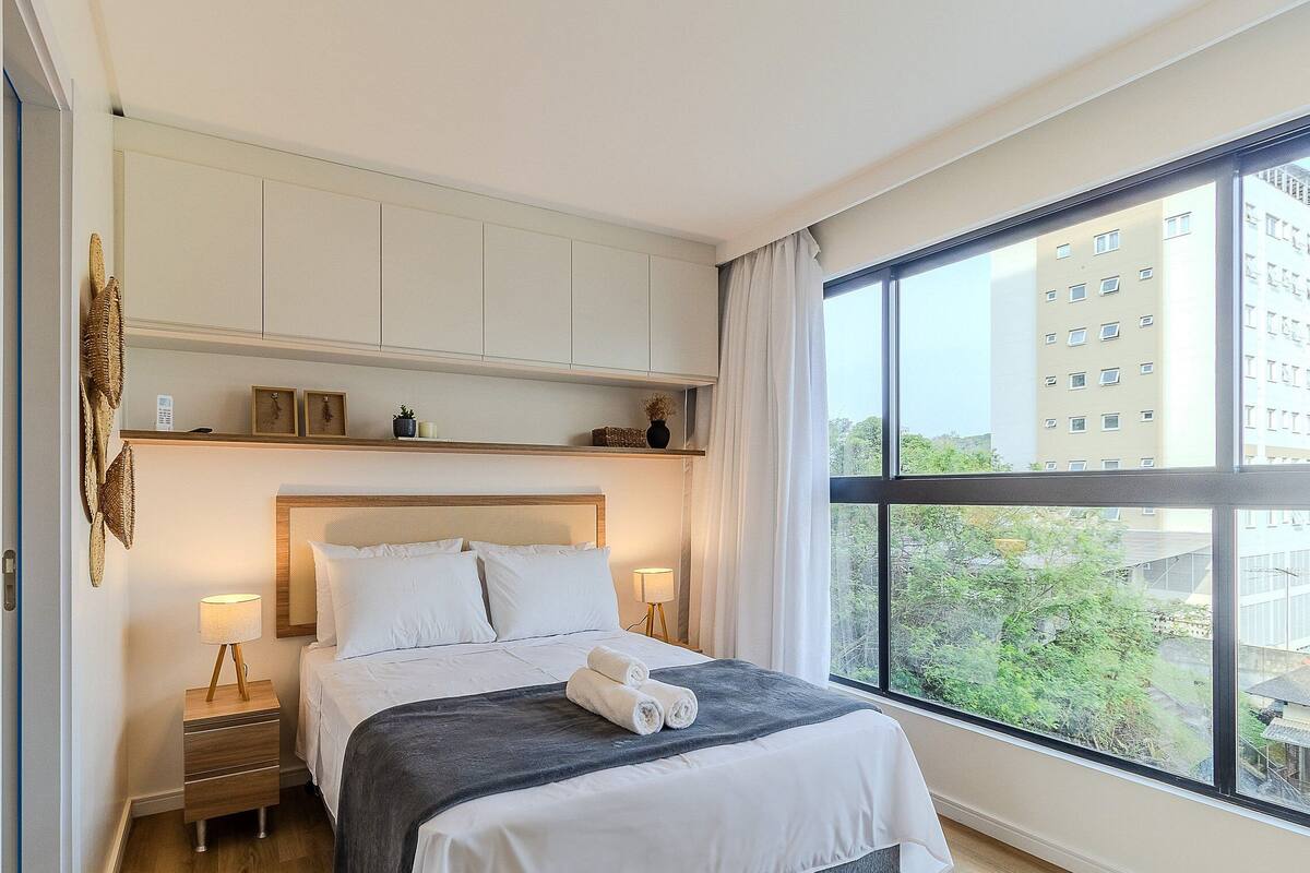 A hotel room with a bed, bedside tables, a window with a view of buildings, and a shelf with decorative items.